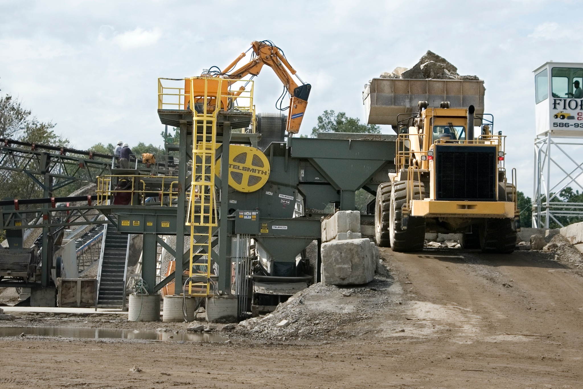 Żółta ładowarka z pełną łyżką kamieni podjeżdża pod stacjonarną kruszarkę Telsmith na terenie zakładu recyklingu materiałów budowlanych, w tle widoczna kabina operatora.