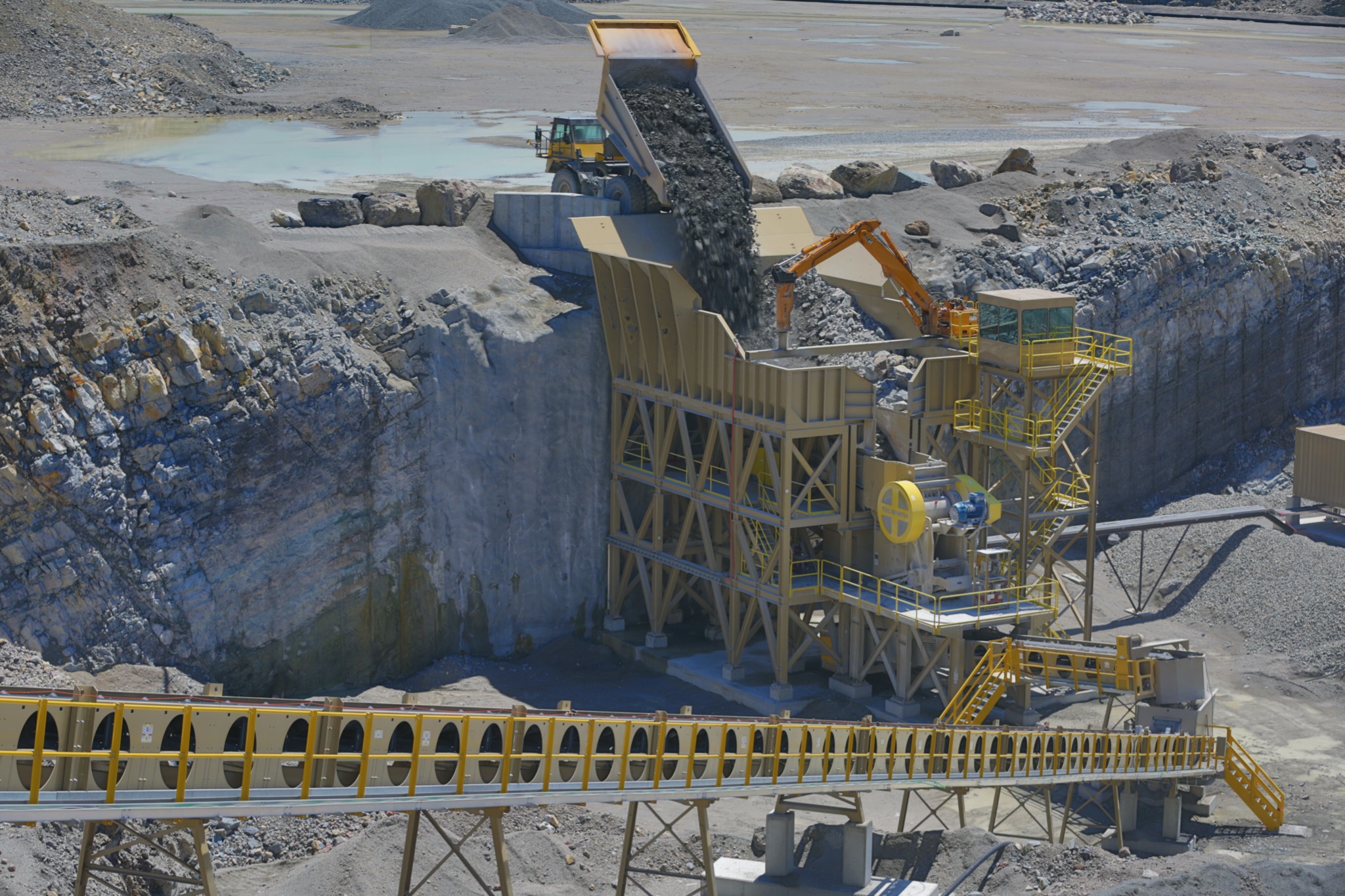 Ciężarówka wyładowuje urobek do kruszarki w kamieniołomie, z widocznym przenośnikiem taśmowym i ścianą skalną w tle.