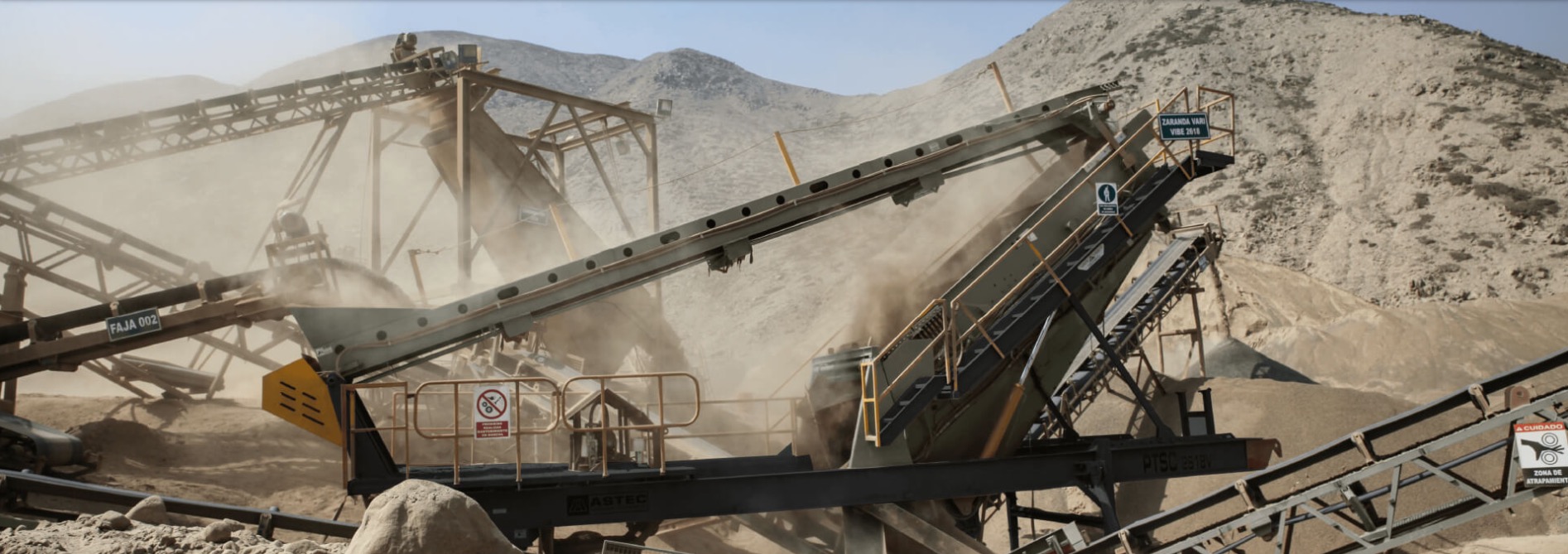 Praca kruszarki do kamienia w kamieniołomie, z widocznymi przenośnikami taśmowymi i pyłem w powietrzu, na tle górzystego krajobrazu.