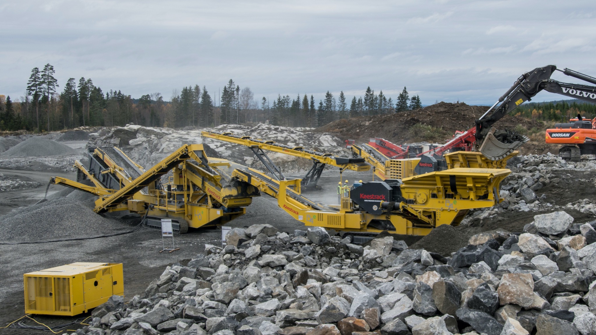 Żółte mobilne przesiewacze i kruszarki Keestrack na placu budowy, z koparką Volvo podającą urobek, w tle las i hałdy kamieni.