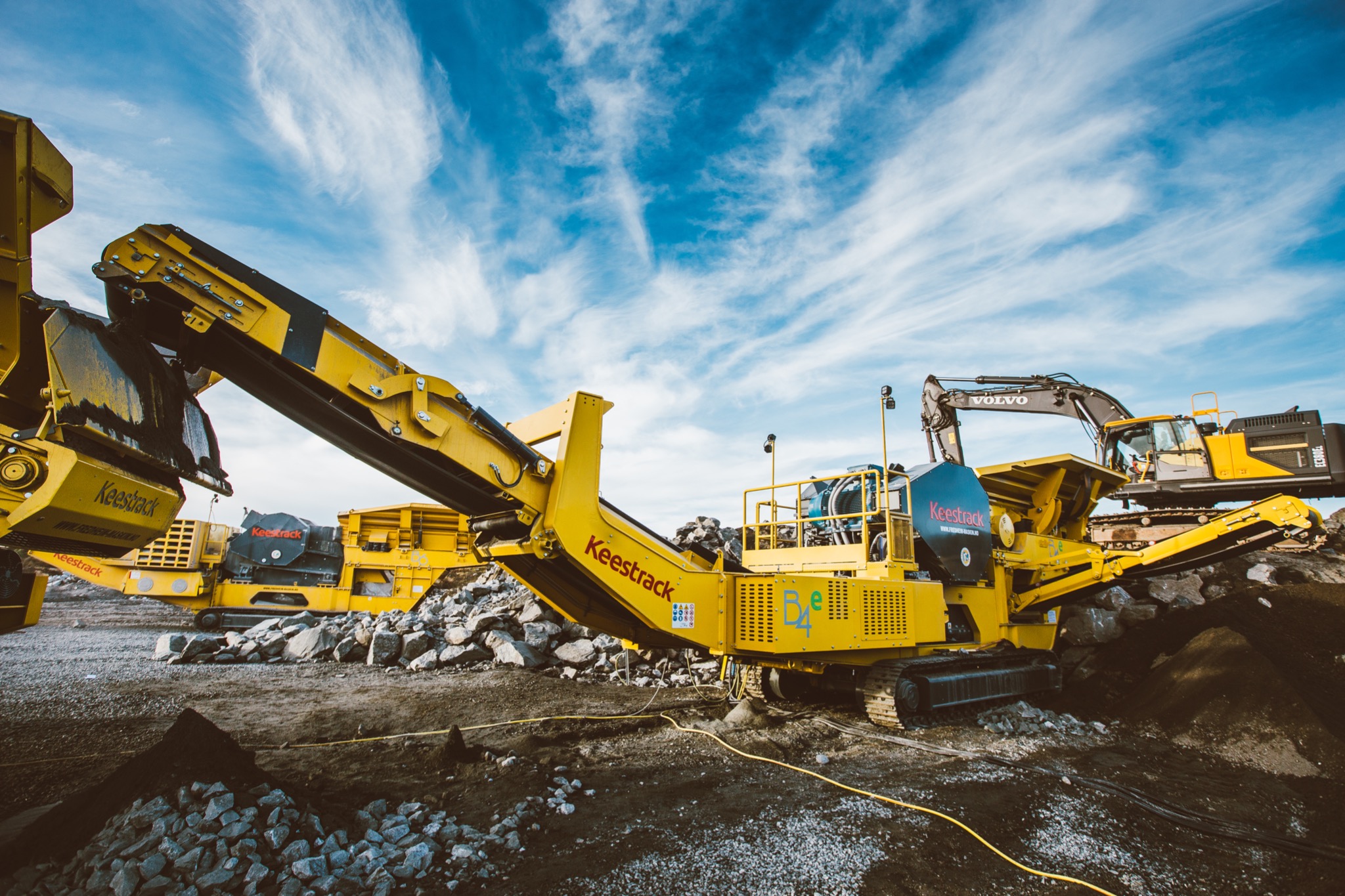 Żółte kruszarki do kamienia marki Keestrack na placu budowy, z koparką Volvo w tle i błękitnym niebem z chmurami.