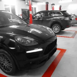 Częściowo odbarwione zdjęcie warsztatu samochodowego z trzema samochodami, w tym czarnym Porsche Cayenne na pierwszym planie, stojącymi na stanowiskach oznaczonych czerwonymi liniami