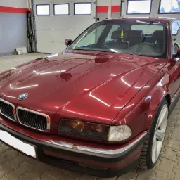 Błyszczący, bordowy samochód BMW serii 7 (E38) po polerowaniu lakieru, zaparkowany w warsztacie z widocznym odświeżaczem powietrza w kształcie choinki na lusterku wstecznym.