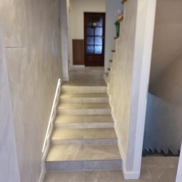 MIAREM - Klatka schodowa z szarymi marmurowymi płytkami na podłodze i schodach, oświetlenie LED wzdłuż ściany, ściany wykończone szarą dekoracyjną fakturą, drewniane drzwi w tle.