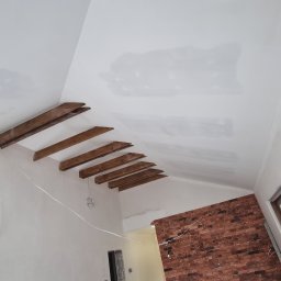 MIAREM - Sufit pod skosem z drewnianymi belkami dekoracyjnymi i fragmentem ściany z czerwonej cegły, częściowo pokryty gładzią, w trakcie prac wykończeniowych.