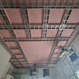 MIAREM - Montaż sufitu podwieszanego z płyt kartonowo-gipsowych na metalowej konstrukcji, widoczne różowe płyty izolacyjne i rusztowanie.
