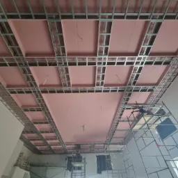 Montaż sufitu podwieszanego z płyt kartonowo-gipsowych na metalowej konstrukcji, widoczne różowe płyty izolacyjne i rusztowanie.