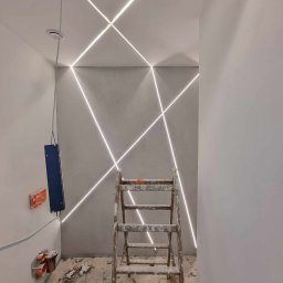 MIAREM - Nowoczesne oświetlenie LED wpuszczone w sufit i ścianę, tworzące geometryczny wzór krzyżujących się linii świetlnych, drabina malarska stoi w pokoju w trakcie remontu, widoczne puszki elektryczne...