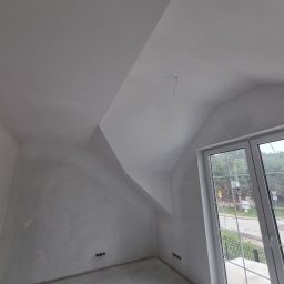 MIAREM - Poddasze ze skośnym sufitem, ściany przygotowane pod malowanie, widoczne gniazdka elektryczne i okno z widokiem na zewnątrz.