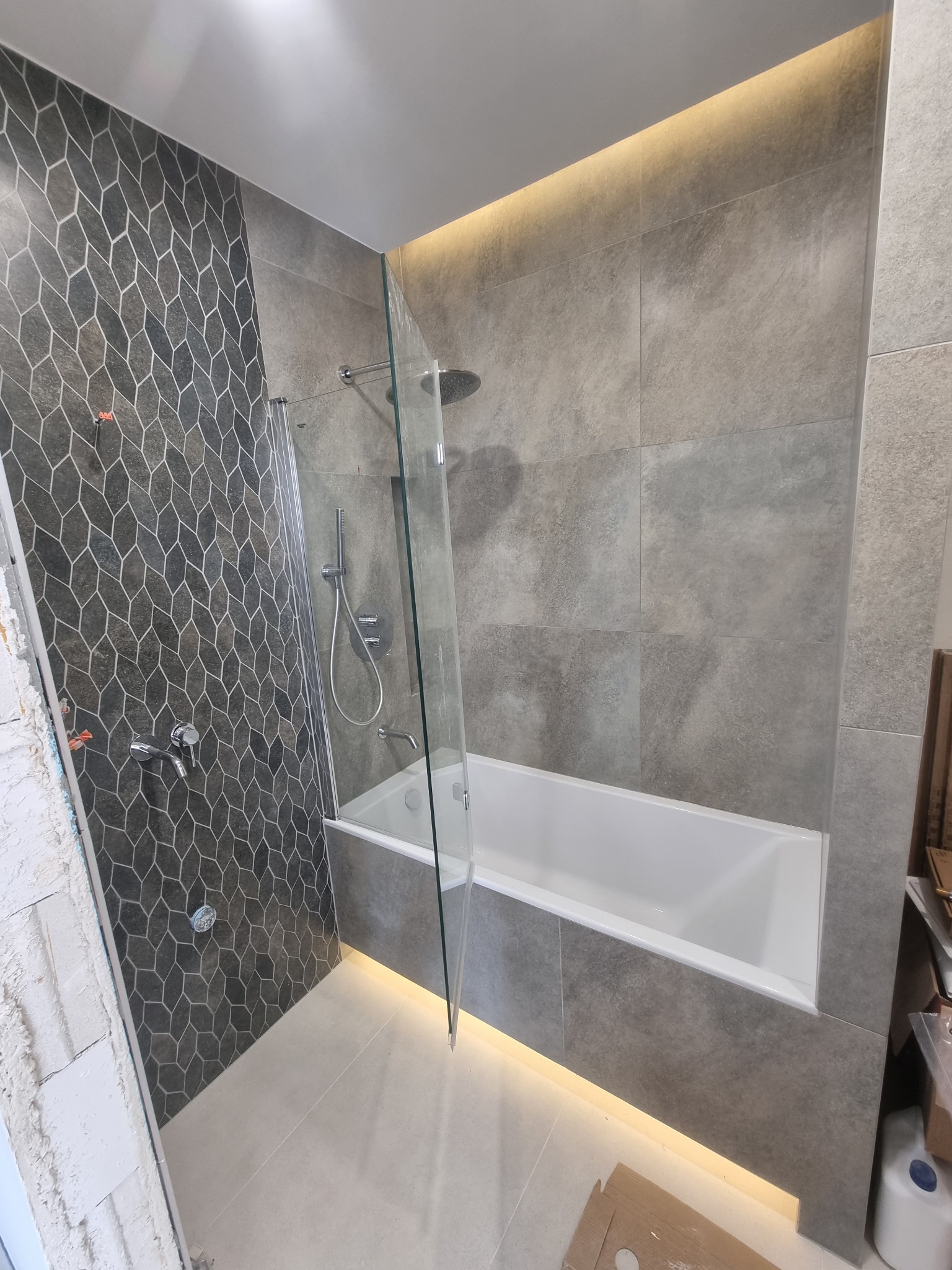 Wykończona łazienka z wanną obudowaną szarymi płytkami, szklanym parawanem nawannowym i mozaiką na ścianie, z oświetleniem LED pod wanną i na suficie.