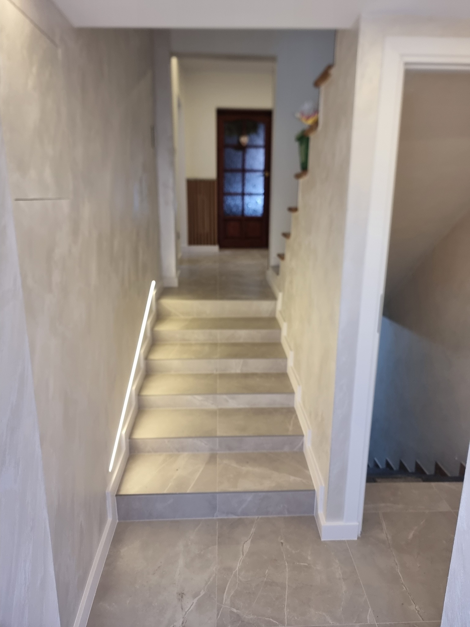 Klatka schodowa z szarymi marmurowymi płytkami na podłodze i schodach, oświetlenie LED wzdłuż ściany, ściany wykończone szarą dekoracyjną fakturą, drewniane drzwi w tle.