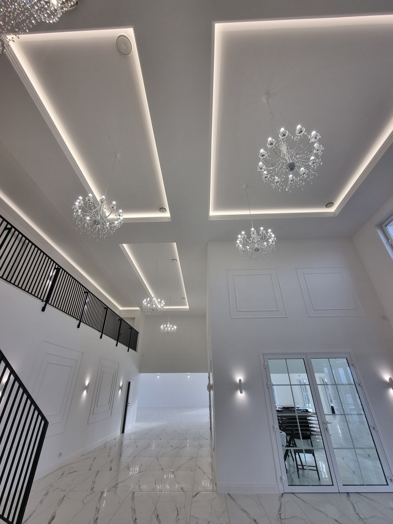 Wnętrze przestronnego, dwukondygnacyjnego holu z marmurową posadzką, białymi ścianami z dekoracyjnymi panelami, czarną balustradą schodów i sufitami z oświetleniem LED oraz kryształowymi żyrandolami.