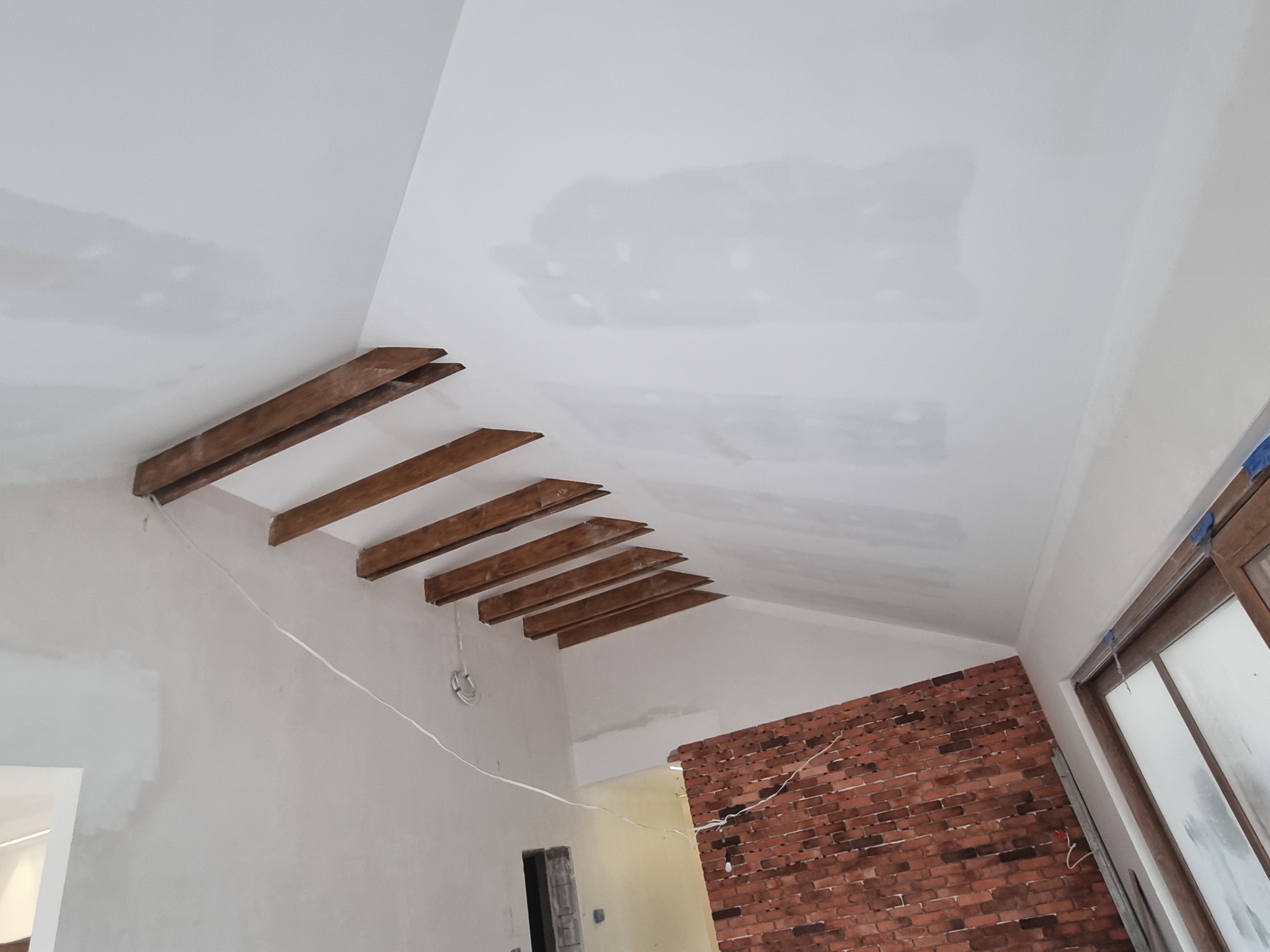 Sufit pod skosem z drewnianymi belkami dekoracyjnymi i fragmentem ściany z czerwonej cegły, częściowo pokryty gładzią, w trakcie prac wykończeniowych.