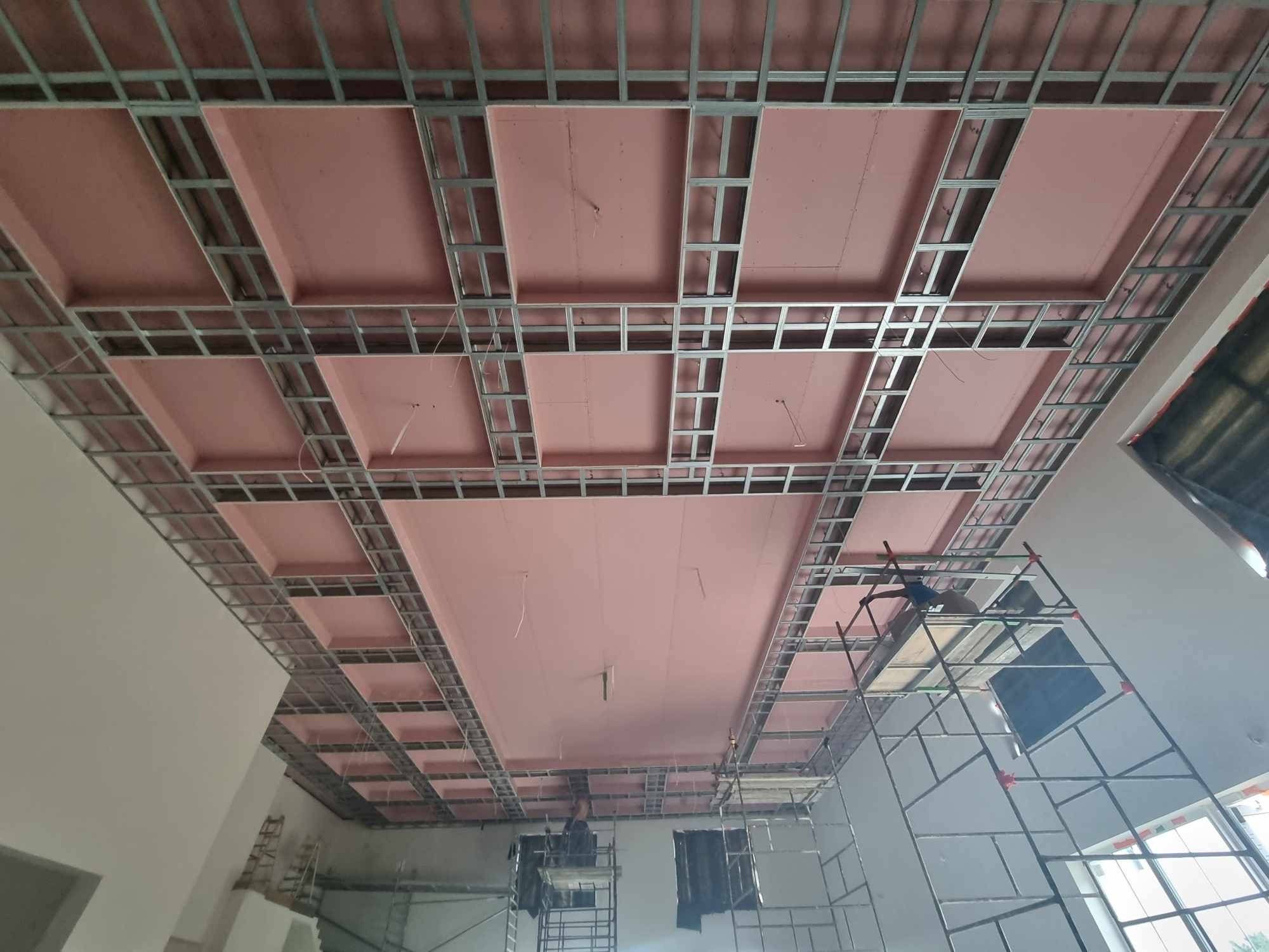 Montaż sufitu podwieszanego z płyt kartonowo-gipsowych na metalowej konstrukcji, widoczne różowe płyty izolacyjne i rusztowanie.
