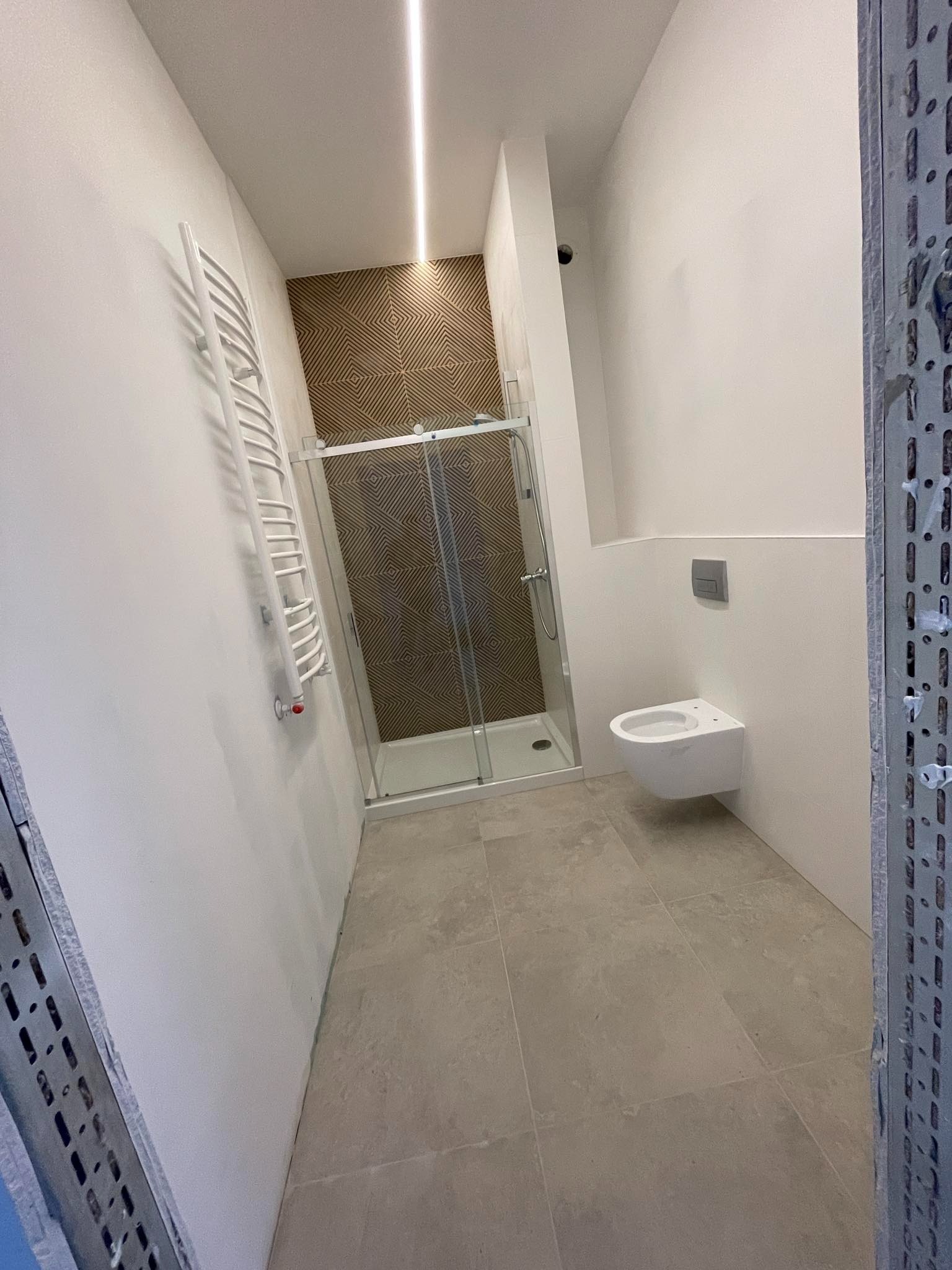 Nowoczesna łazienka w trakcie wykańczania: kabina prysznicowa z geometrycznym wzorem na ścianie, podwieszana toaleta, grzejnik drabinkowy i surowy metalowy profil na pierwszym planie.