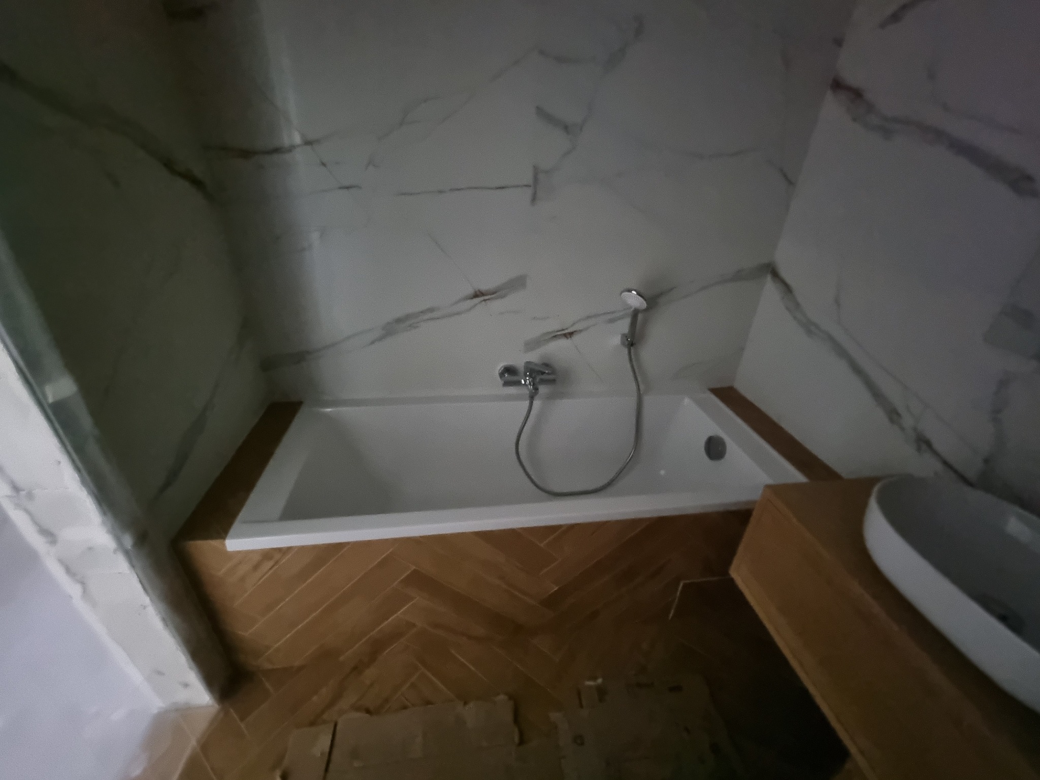 Biała wanna z zamontowaną baterią i słuchawką prysznicową, obudowana drewnopodobną płytką ułożoną w jodełkę, ściany wykończone marmurową płytką, widoczna umywalka nablatowa.