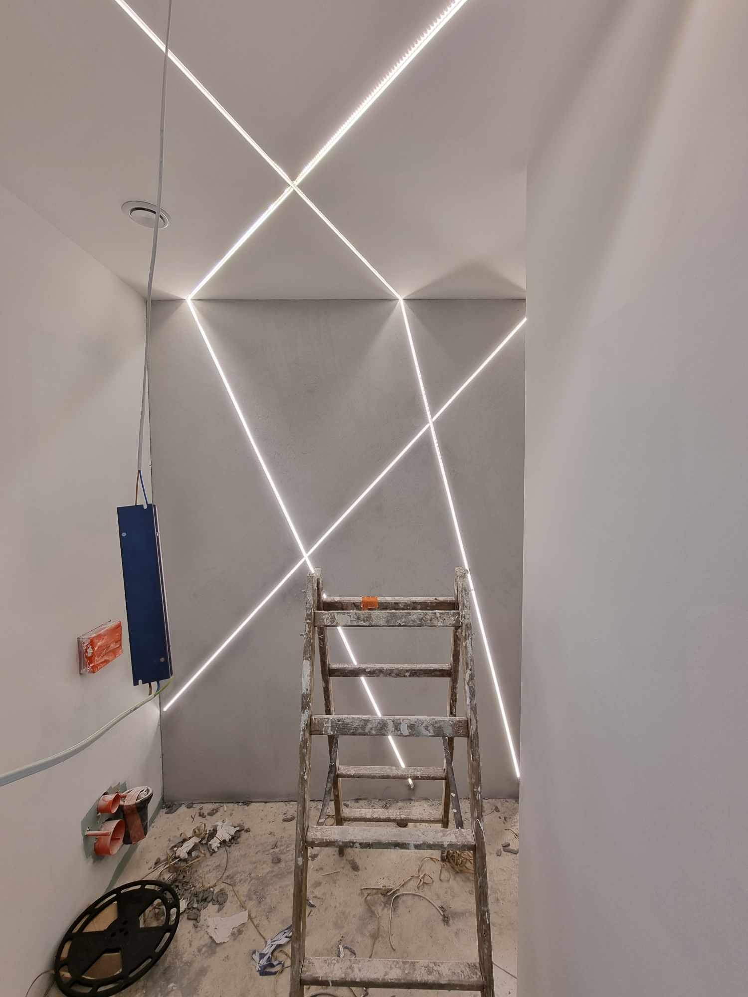 Nowoczesne oświetlenie LED wpuszczone w sufit i ścianę, tworzące geometryczny wzór krzyżujących się linii świetlnych, drabina malarska stoi w pokoju w trakcie remontu, widoczne puszki elektryczne...