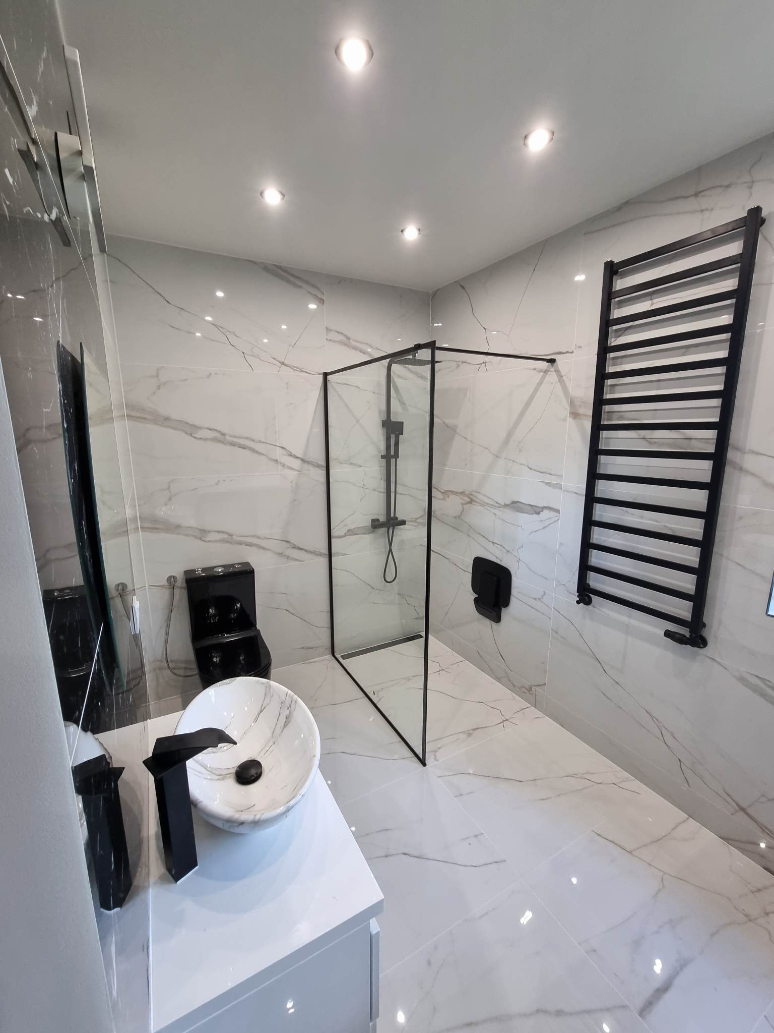 Nowoczesna łazienka wykończona płytkami imitującymi marmur, z czarną armaturą i grzejnikiem drabinkowym, kabina prysznicowa typu walk-in z czarnymi profilami.