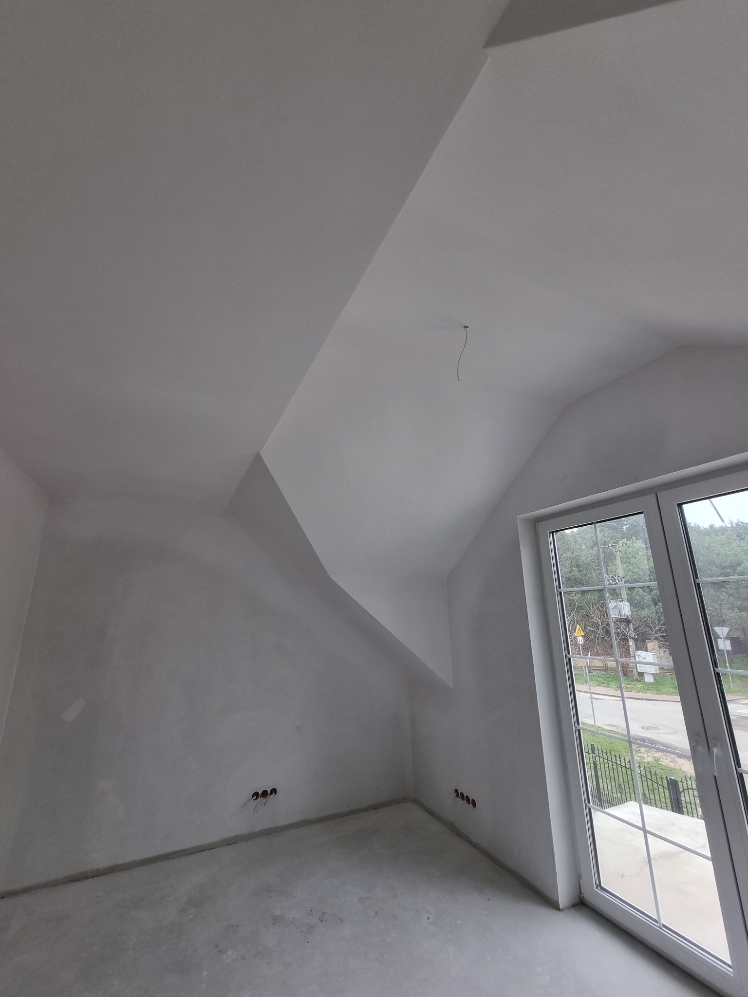 Poddasze ze skośnym sufitem, ściany przygotowane pod malowanie, widoczne gniazdka elektryczne i okno z widokiem na zewnątrz.