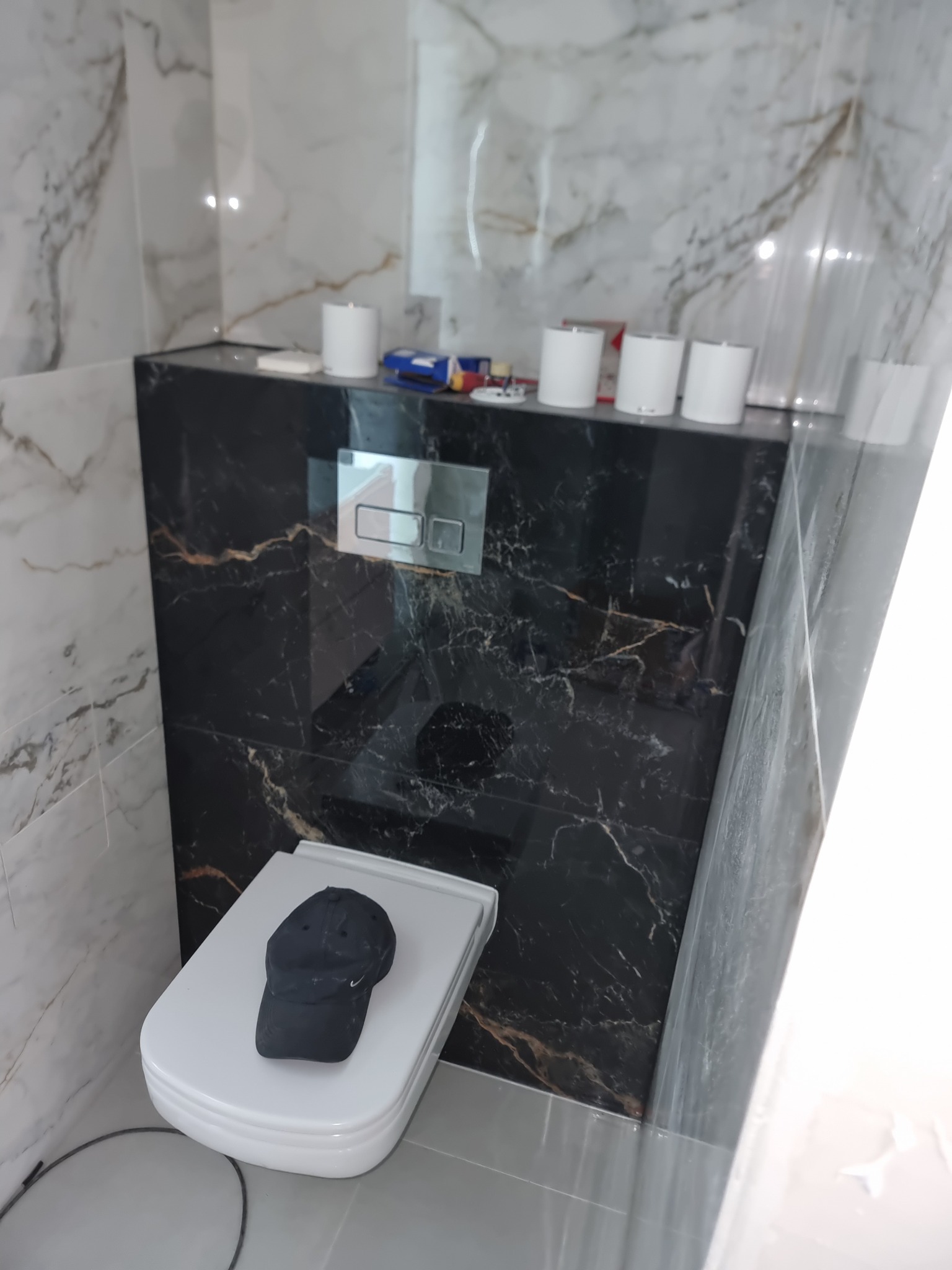 Zabudowa stelaża WC z czarnego marmuru z widocznym przyciskiem spłukującym i czapką na desce sedesowej, wykończona płytkami imitującymi biały marmur na ścianach.