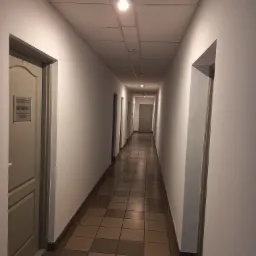 Długi korytarz z białymi ścianami i brązowo-beżową podłogą z płytek, oświetlony przez lampy sufitowe, z drzwiami po obu stronach, na jednych z nich tabliczka informacyjna 'METROSAT'.