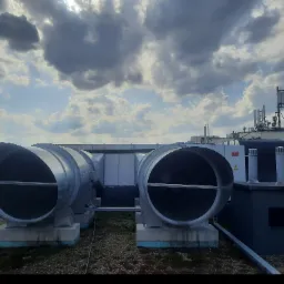 Dwa duże, metalowe wloty wentylacyjne na dachu budynku, z widocznymi elementami instalacji i tłem błękitnego nieba z chmurami.