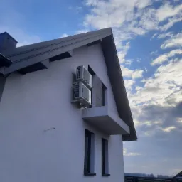 Dwa klimatyzatory zamontowane na elewacji nowoczesnego domu jednorodzinnego z balkonem, na tle błękitnego nieba z obłokami.