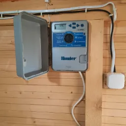 Panel sterowania systemem nawadniania Hunter X-Core zamontowany na drewnianej ścianie, z otwartą obudową i widocznymi przewodami zasilającymi.