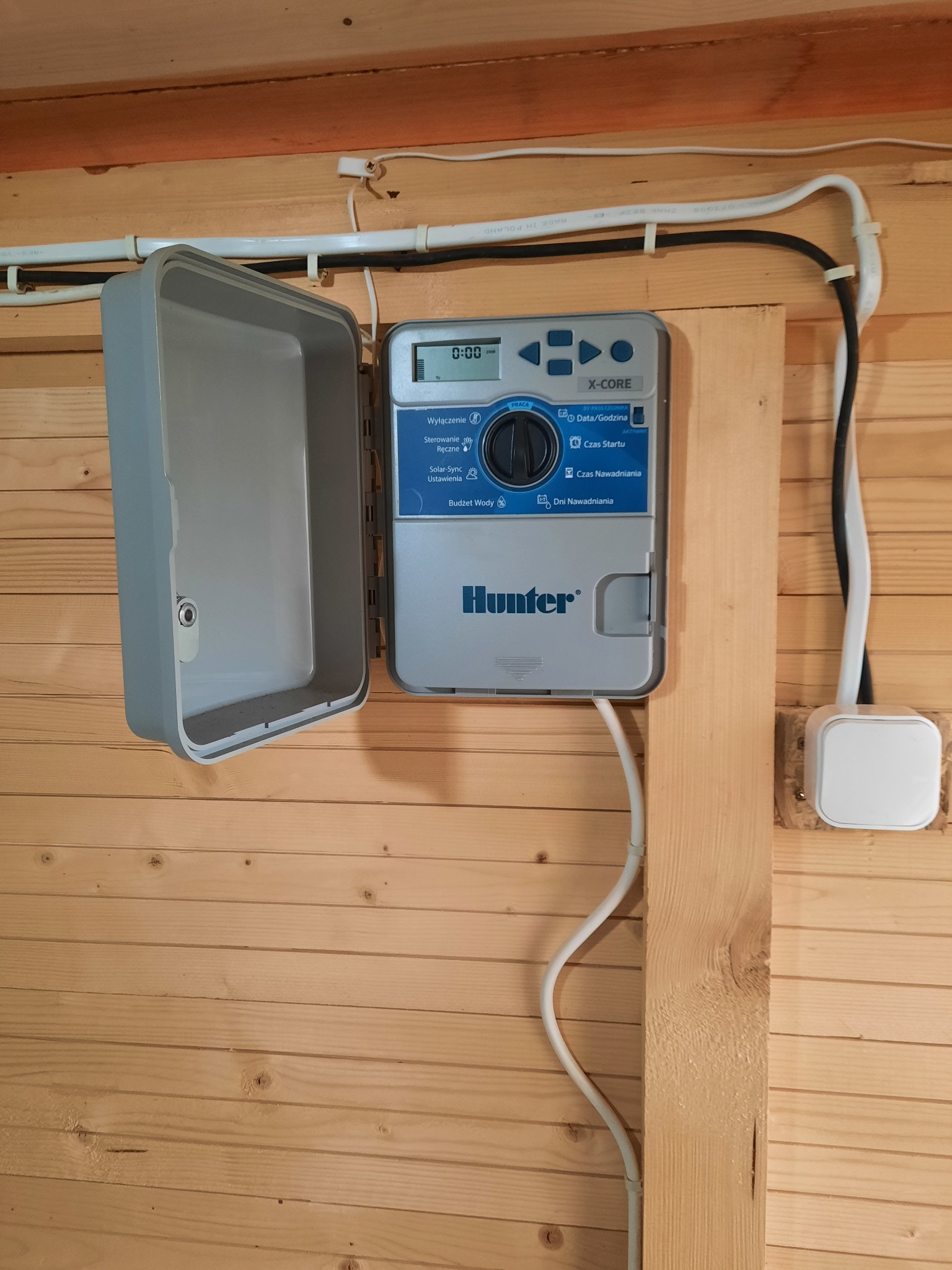 Panel sterowania systemem nawadniania Hunter X-Core zamontowany na drewnianej ścianie, z otwartą obudową i widocznymi przewodami zasilającymi.