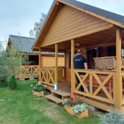 Dwa drewniane domki letniskowe z werandami, przed jednym stoi osoba w czarnej koszulce z logo, na werandzie krzesło wiklinowe, na schodach butelka z rozpylaczem, kwiaty w drewnianych skrzynkach...