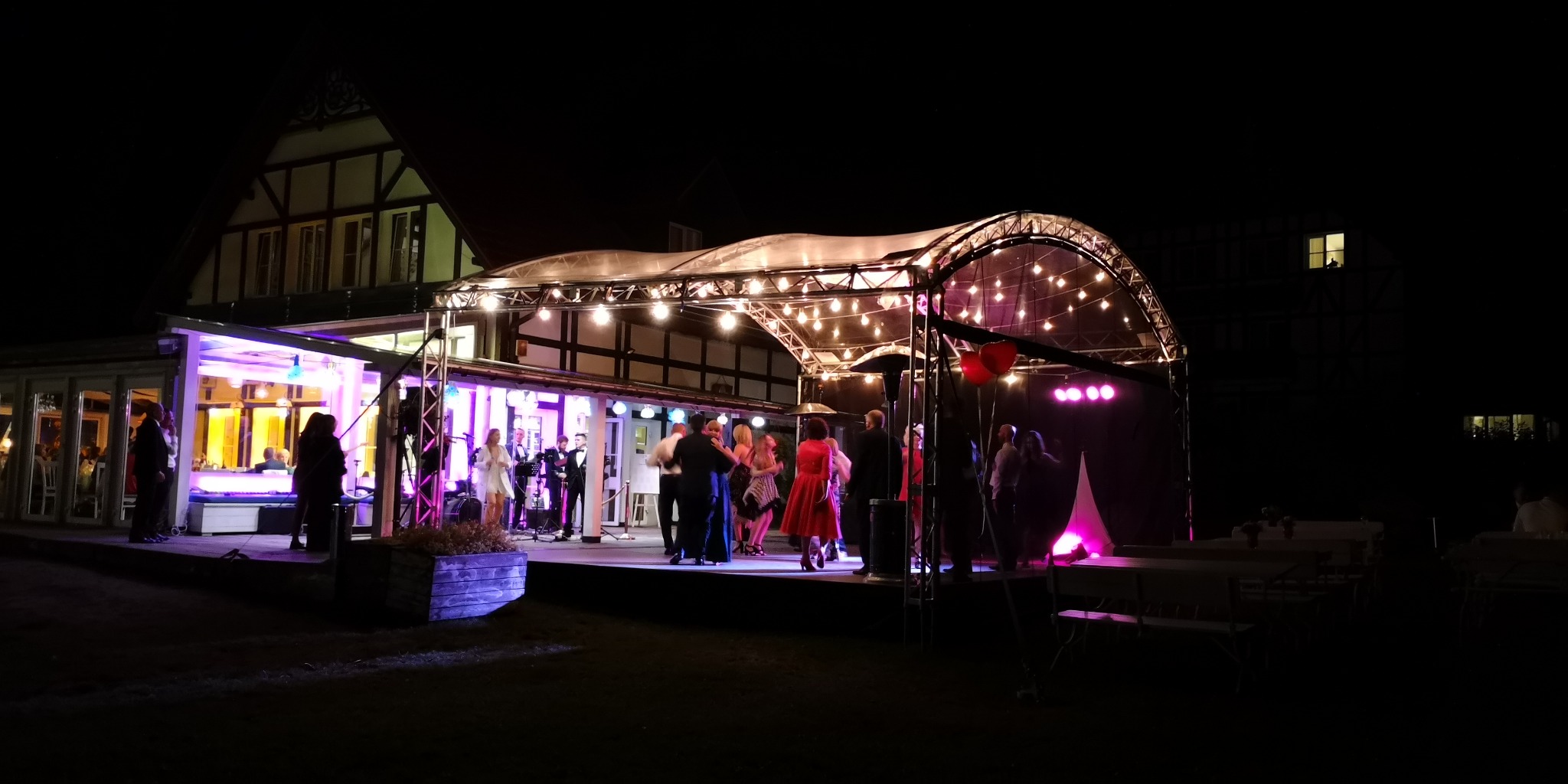 Wieczorne przyjęcie z zespołem na scenie oświetlonej girlandami żarówek i purpurowymi reflektorami. W tle budynek z elementami muru pruskiego.