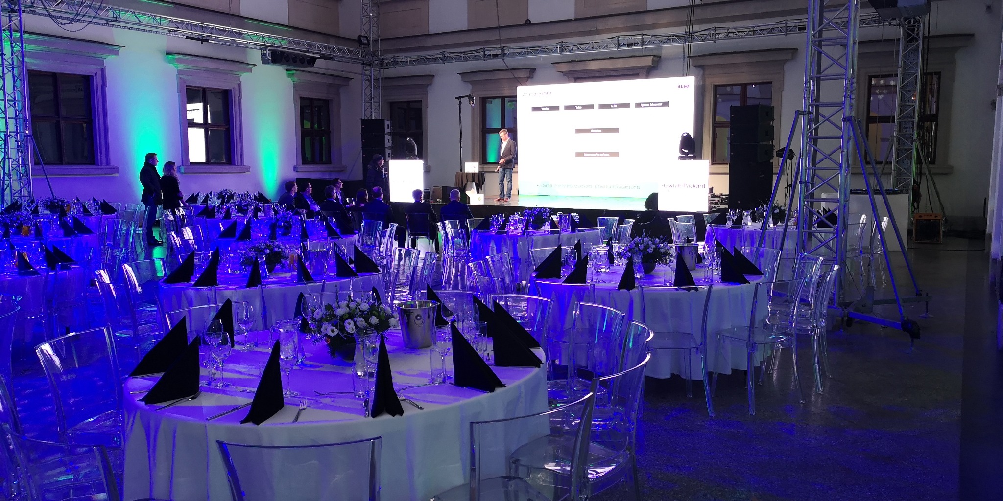 Sala bankietowa z okrągłymi stołami nakrytymi białymi obrusami i czarnymi serwetkami, transparentne krzesła, scena z prelegentem i ekranem, niebieskie oświetlenie.