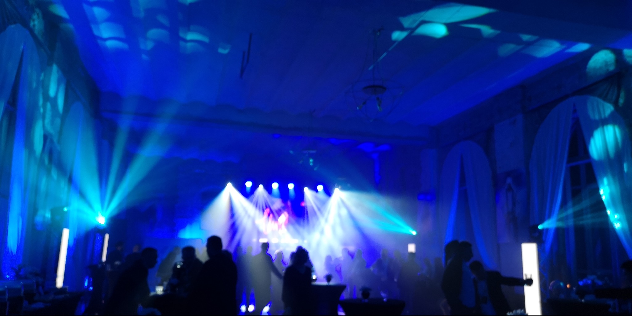 Sala balowa oświetlona niebieskimi reflektorami podczas wydarzenia, widoczni goście w tle, dekoracje okienne z białych tkanin.