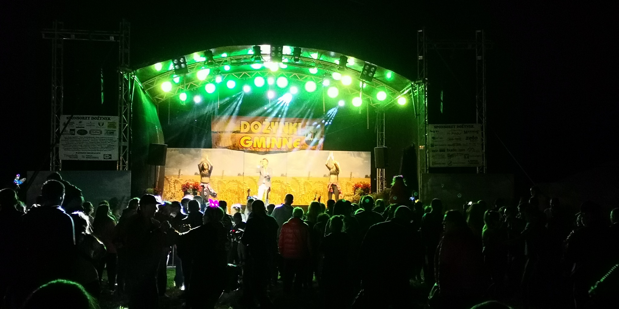 Scena plenerowa oświetlona zielonymi reflektorami podczas dożynek gminnych, z występującymi tancerkami i piosenkarzem na tle dekoracji przedstawiającej pole zboża, widoczny tłum publiczności.