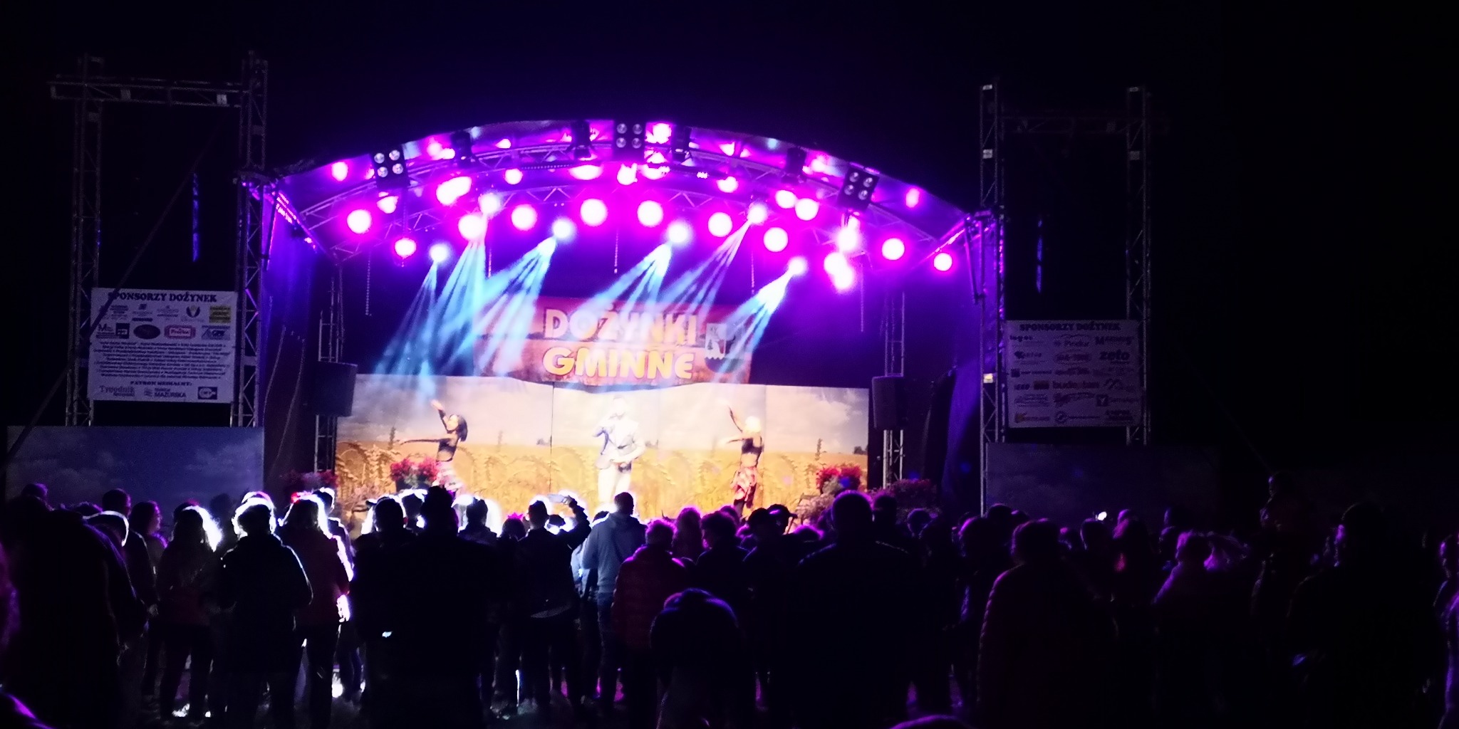 Scena oświetlona fioletowymi reflektorami podczas wieczornego wydarzenia plenerowego w Piszu, z widoczną publicznością i banerami sponsorskimi po bokach sceny.