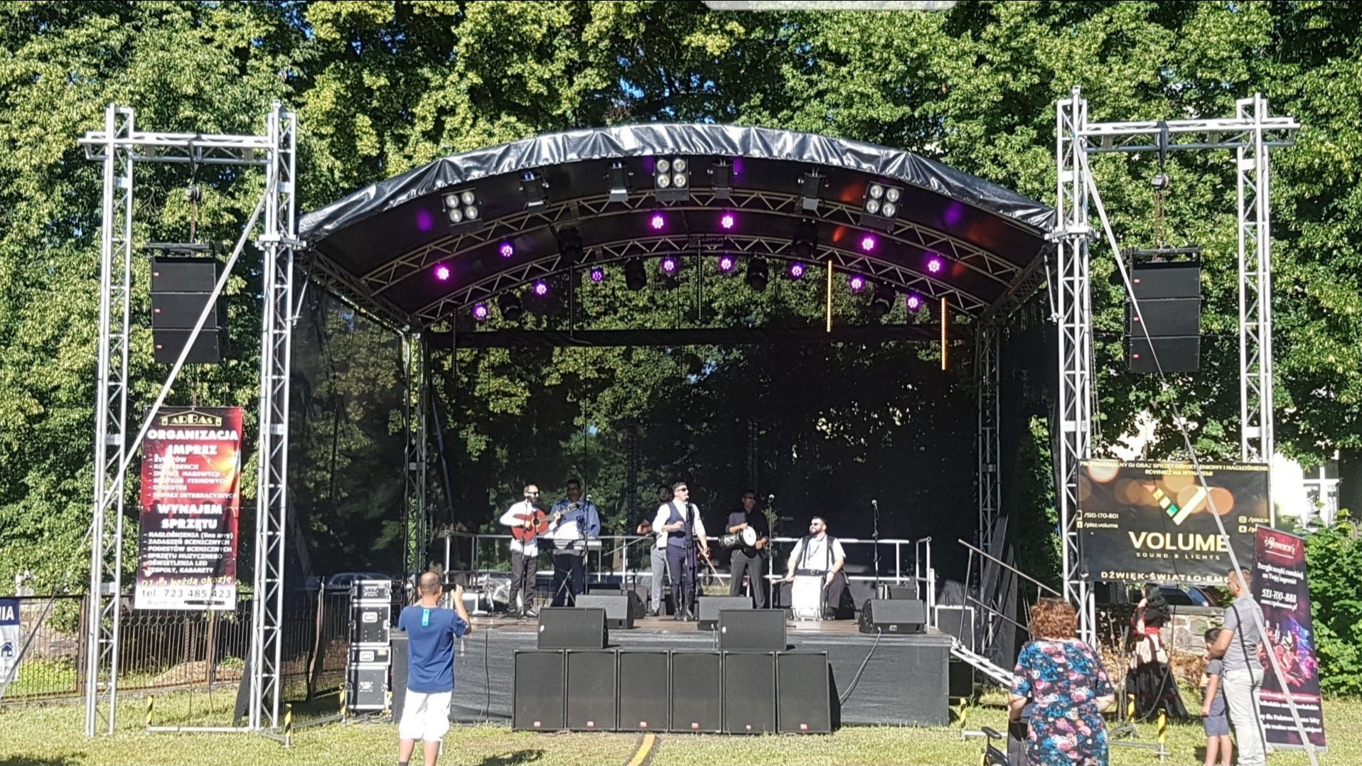 Zespół muzyczny na scenie plenerowej z nagłośnieniem i oświetleniem, widoczni widzowie.