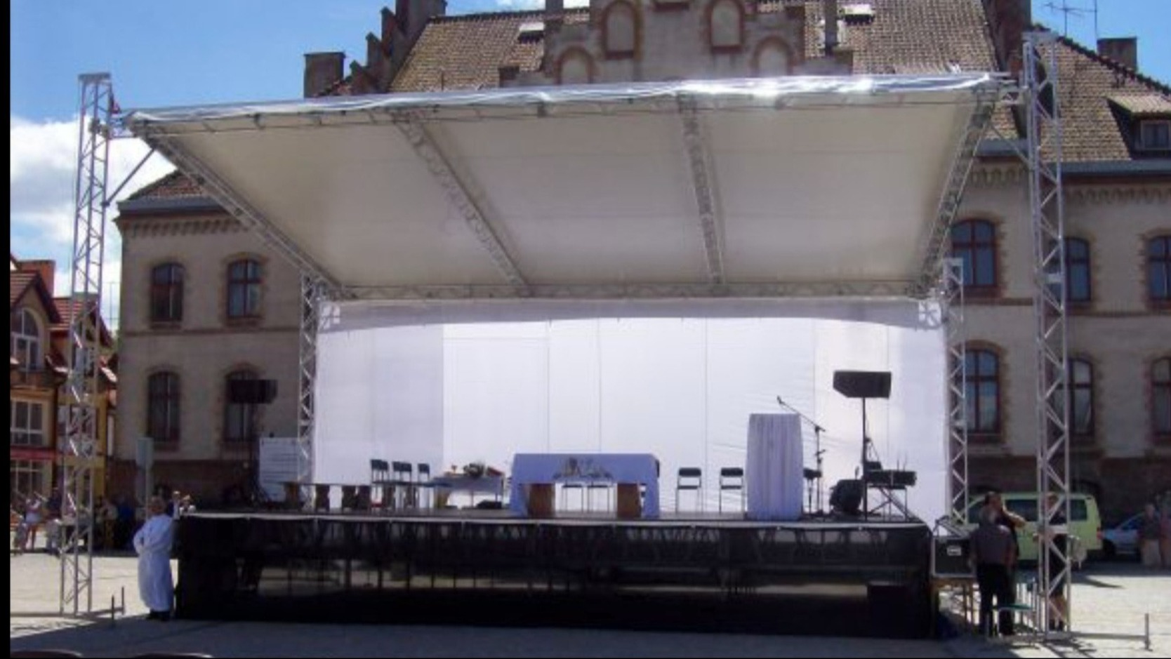 Scena plenerowa z zadaszeniem i białym tłem, przygotowana do uroczystości religijnej na tle budynku z czerwonej cegły.