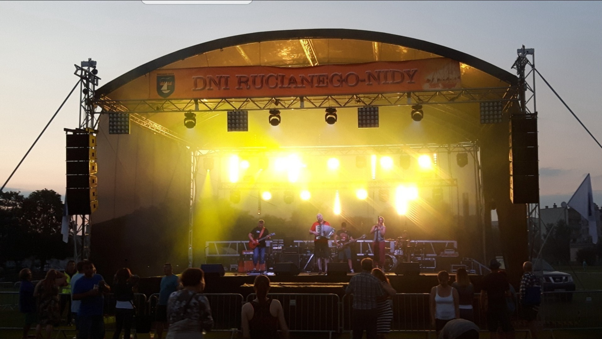 Scena plenerowa oświetlona reflektorami podczas koncertu Dni Ruciane-Nida, zespół muzyczny na scenie, widoczni widzowie przed sceną.