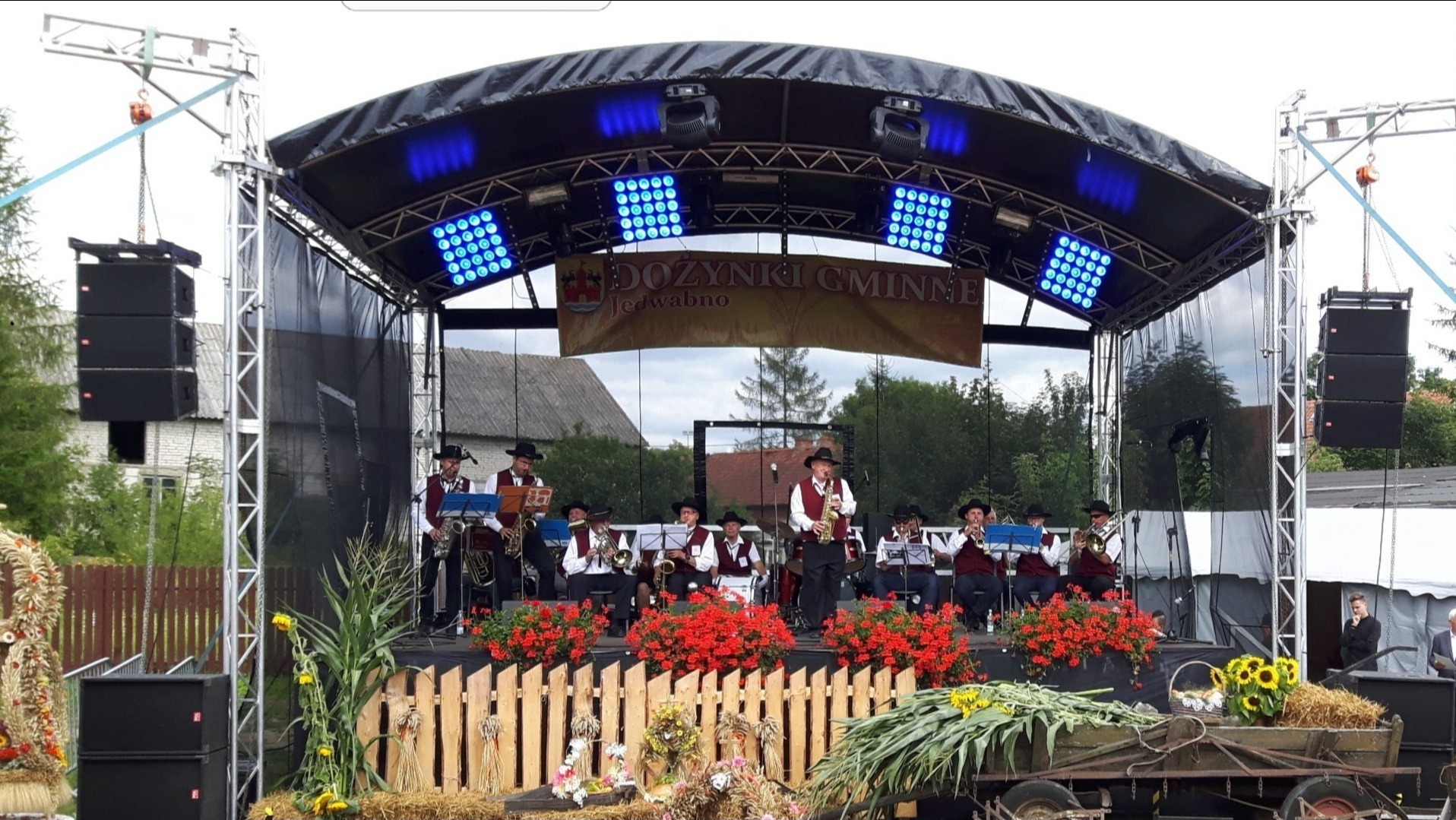 Orkiestra dęta w tradycyjnych strojach występuje na scenie podczas dożynek gminnych, podświetlona niebieskimi reflektorami, z kolumnami głośnikowymi po bokach. Na pierwszym planie dekoracje...