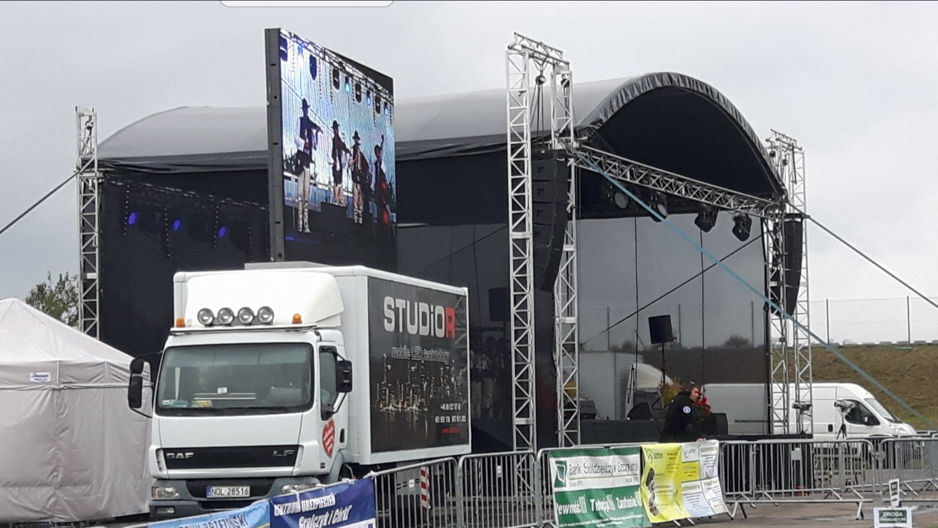 Mobilna scena z dużym ekranem LED wyświetlającym występ zespołu, ustawiona obok ciężarówki z logo 'STUDIO R mobile LED technology', widoczny namiot cateringowy i metalowe barierki.