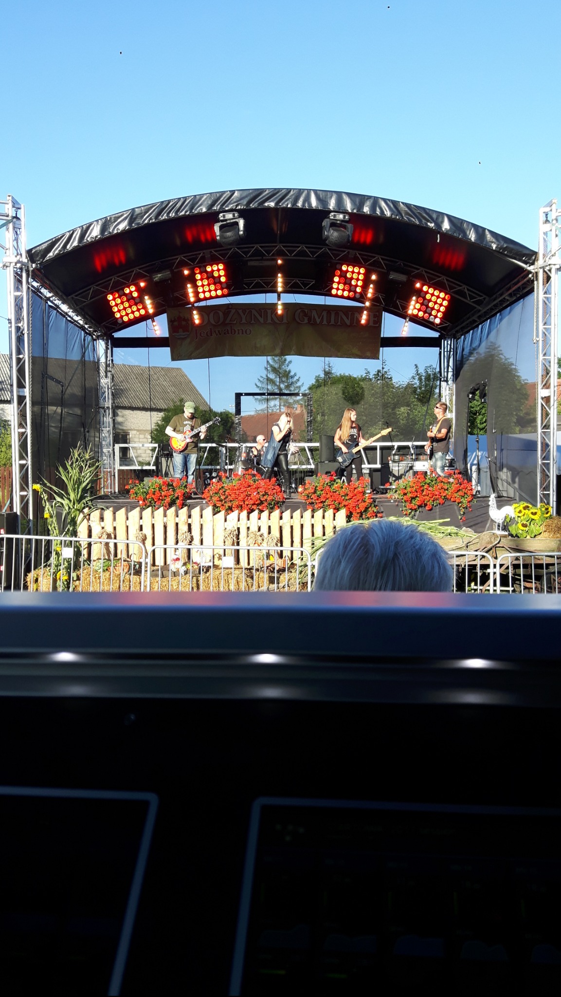 Zespół muzyczny na scenie o wymiarach 6x8 z czerwonymi kwiatami, oświetleniem punktowym i banerem 'Dożynki Gminne Jedwabno' w tle, widok zza kulis.