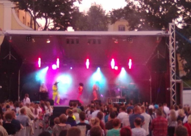 Scena plenerowa z występem artystycznym, oświetlona reflektorami w kolorach niebieskim i różowym, widownia publiczności w tle.