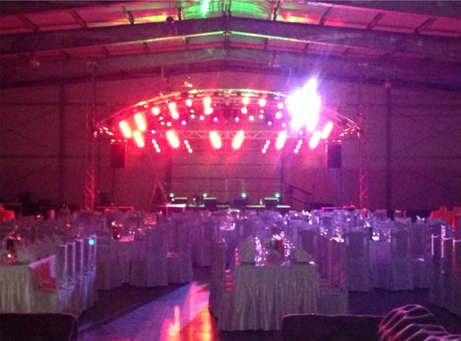 Sala bankietowa z oświetleniem scenicznym, stoły nakryte białymi obrusami, scena z konstrukcją oświetleniową w kolorze różowym i zielonym, przygotowana na imprezę.