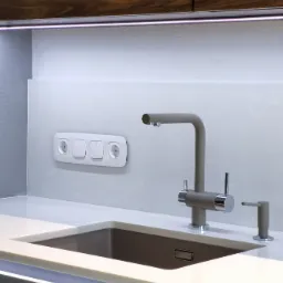 Nowoczesna, minimalistyczna kuchnia: szary kran z dozownikiem mydła obok zlewu wpuszczonego w biały blat, na tle marmurowej ściany z gniazdkami i włącznikami, oświetlenie LED podszafkowe.