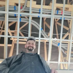 Instalator w roboczym kombinezonie i czapce, patrzący w dół na obiektyw, na tle konstrukcji drewnianej i metalowej z szarymi rurami wentylacyjnymi oznaczonymi niebieską taśmą.