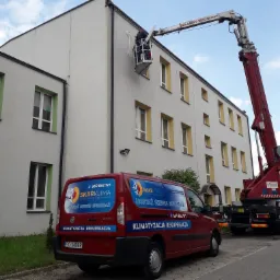 Pracownik na podnośniku koszowym montuje przewody wentylacyjne na elewacji budynku, widoczny firmowy samochód z reklamą usług klimatyzacji i wentylacji.