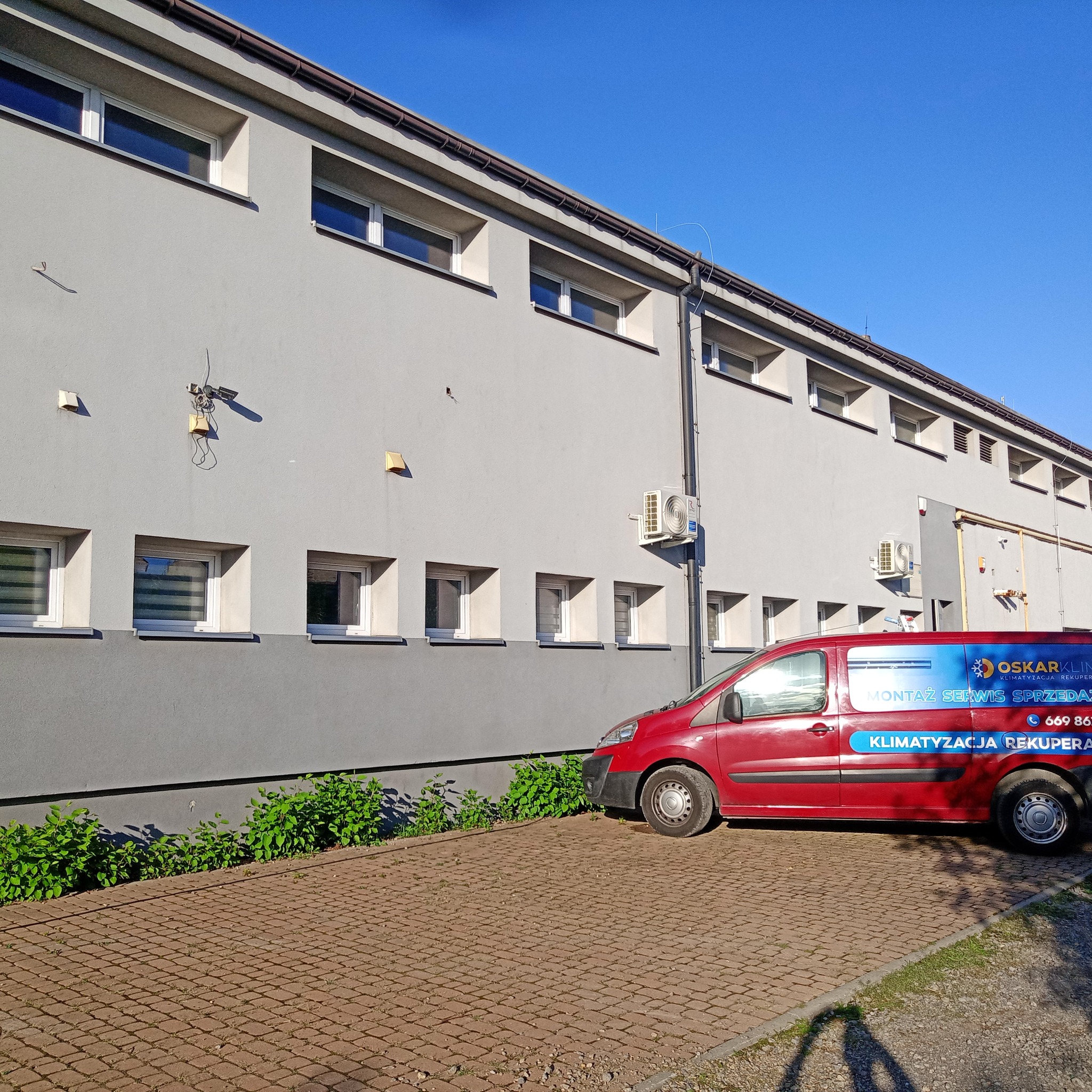 Czerwony samochód serwisowy z logo firmy OskarKlim zaparkowany przed szarym budynkiem z zamontowaną jednostką klimatyzacji na ścianie.