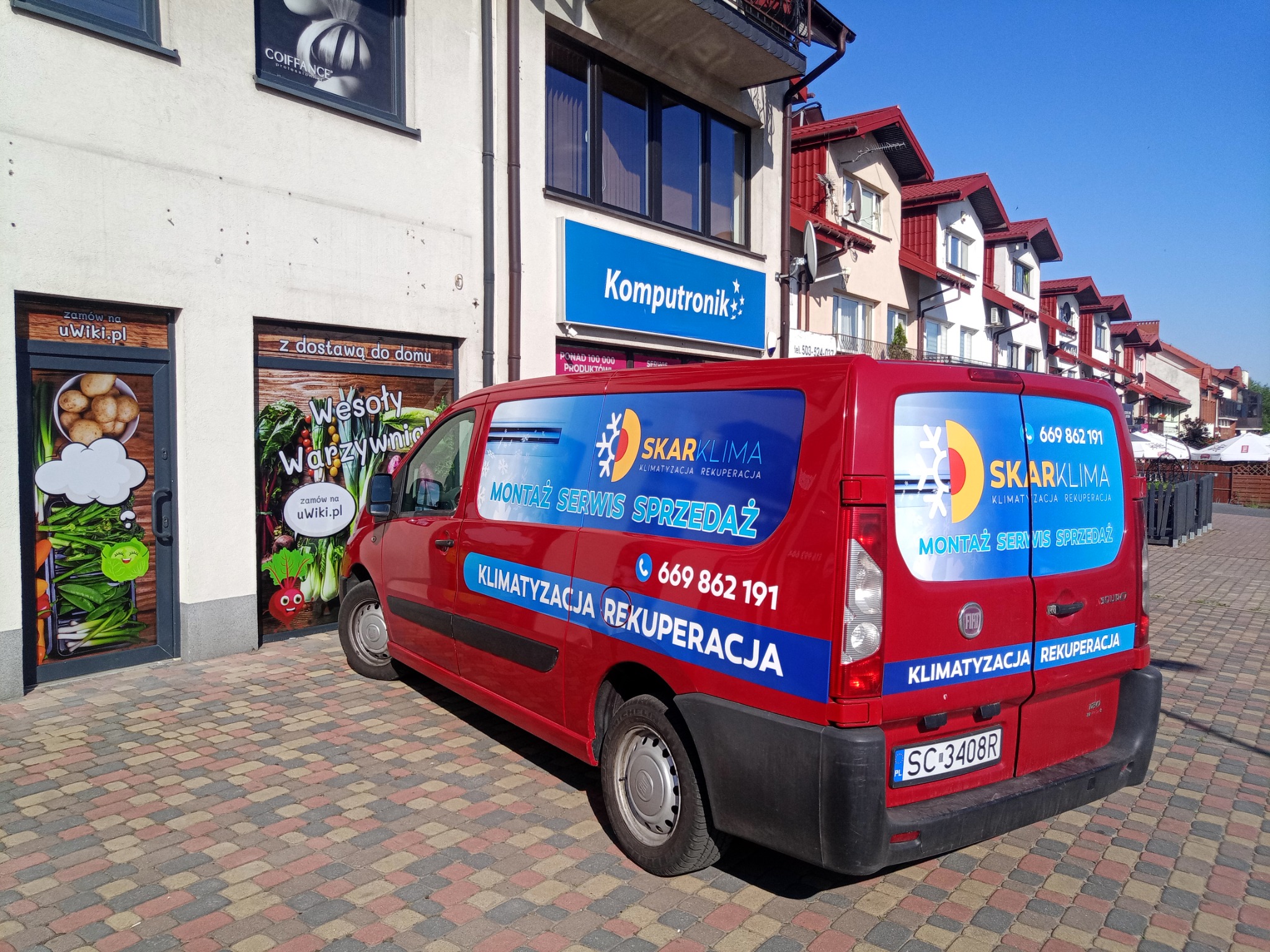 Czerwony samochód dostawczy firmy SkarKlima z reklamą montażu, serwisu i sprzedaży systemów klimatyzacji i rekuperacji, zaparkowany na chodniku z kostki brukowej przed budynkiem z szyldami sklepów.