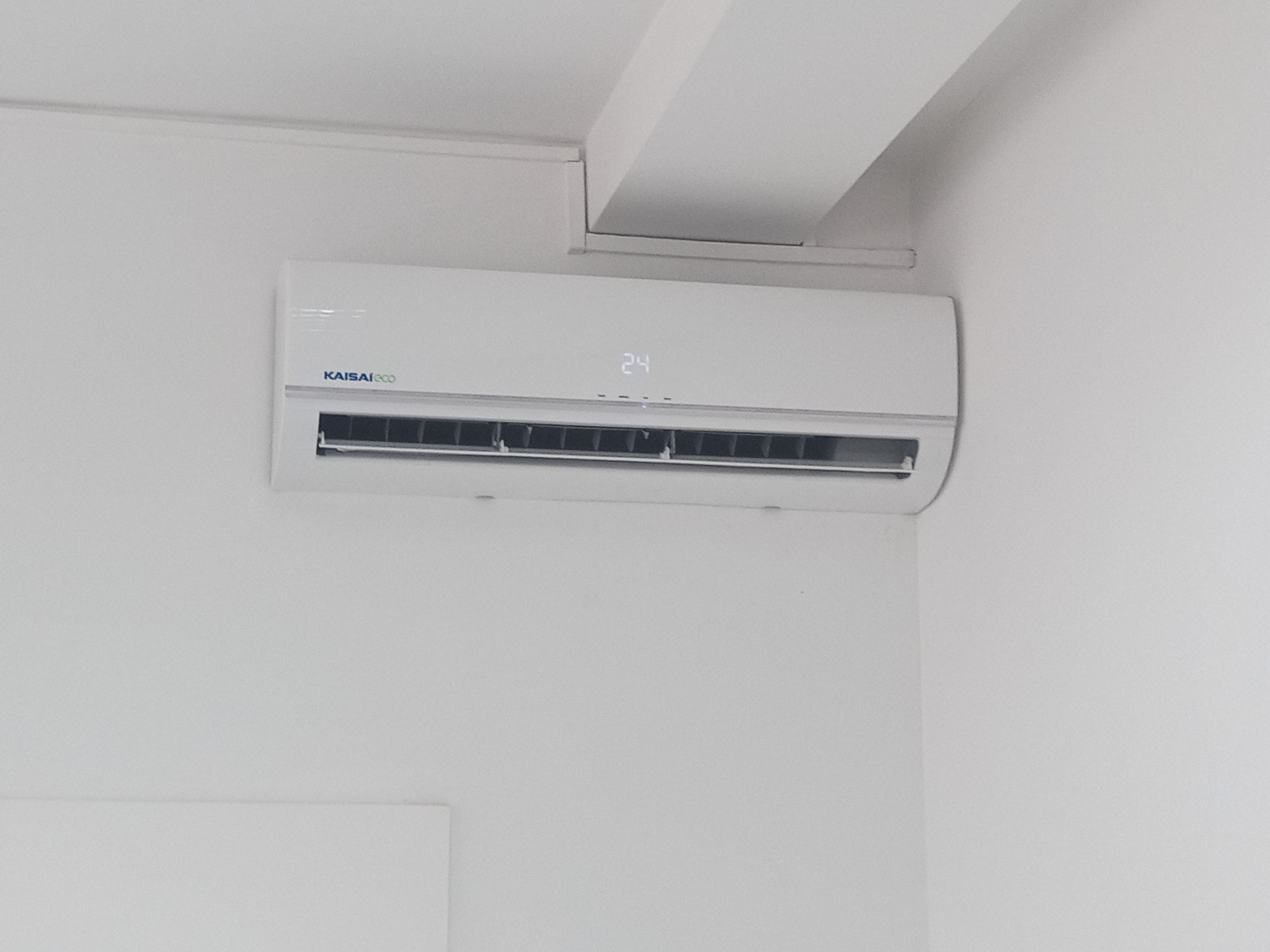 Biały klimatyzator Kaisai Eco z wyświetlaną temperaturą 24 stopnie Celsjusza, zamontowany na białej ścianie w rogu pomieszczenia.