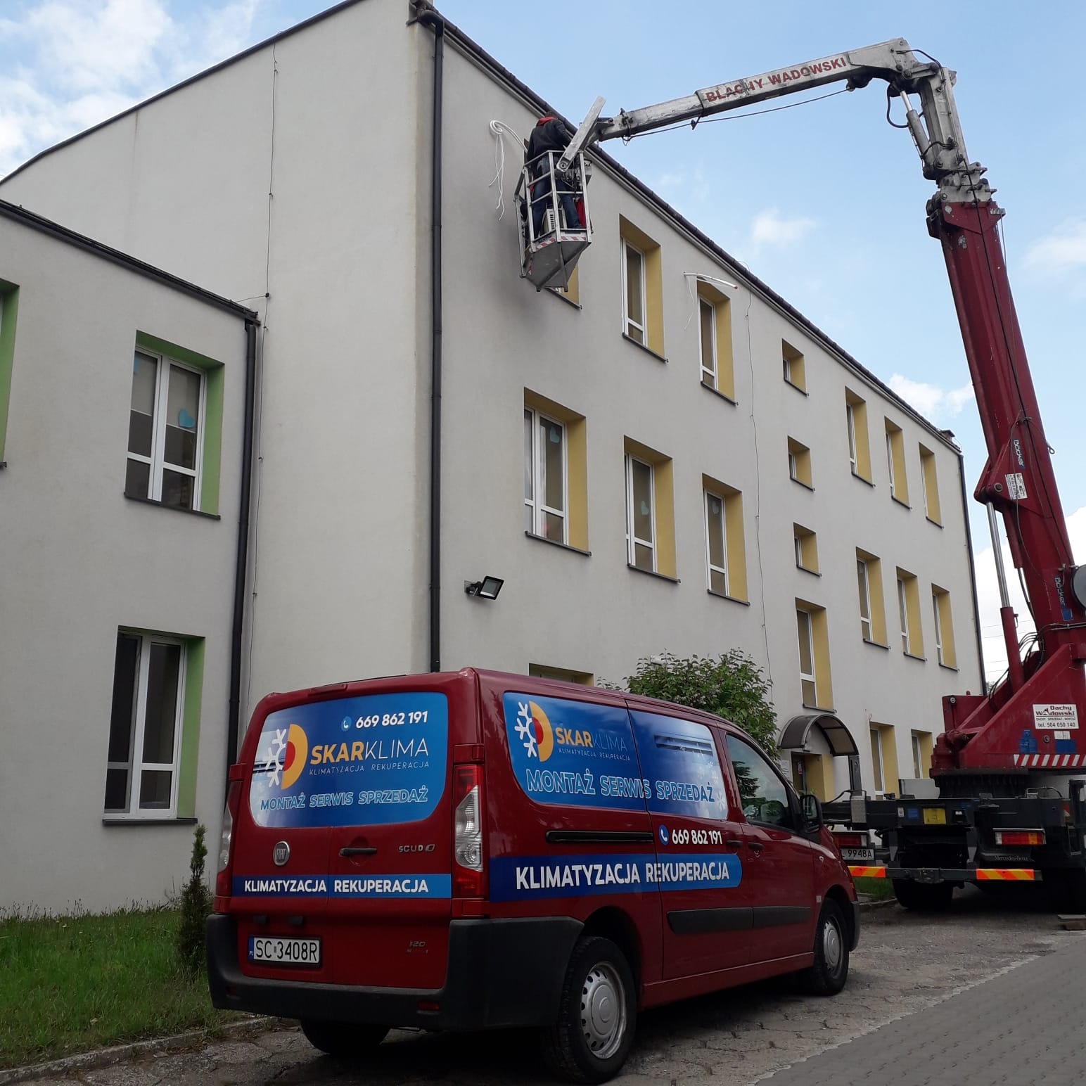 Pracownik na podnośniku koszowym montuje przewody wentylacyjne na elewacji budynku, widoczny firmowy samochód z reklamą usług klimatyzacji i wentylacji.